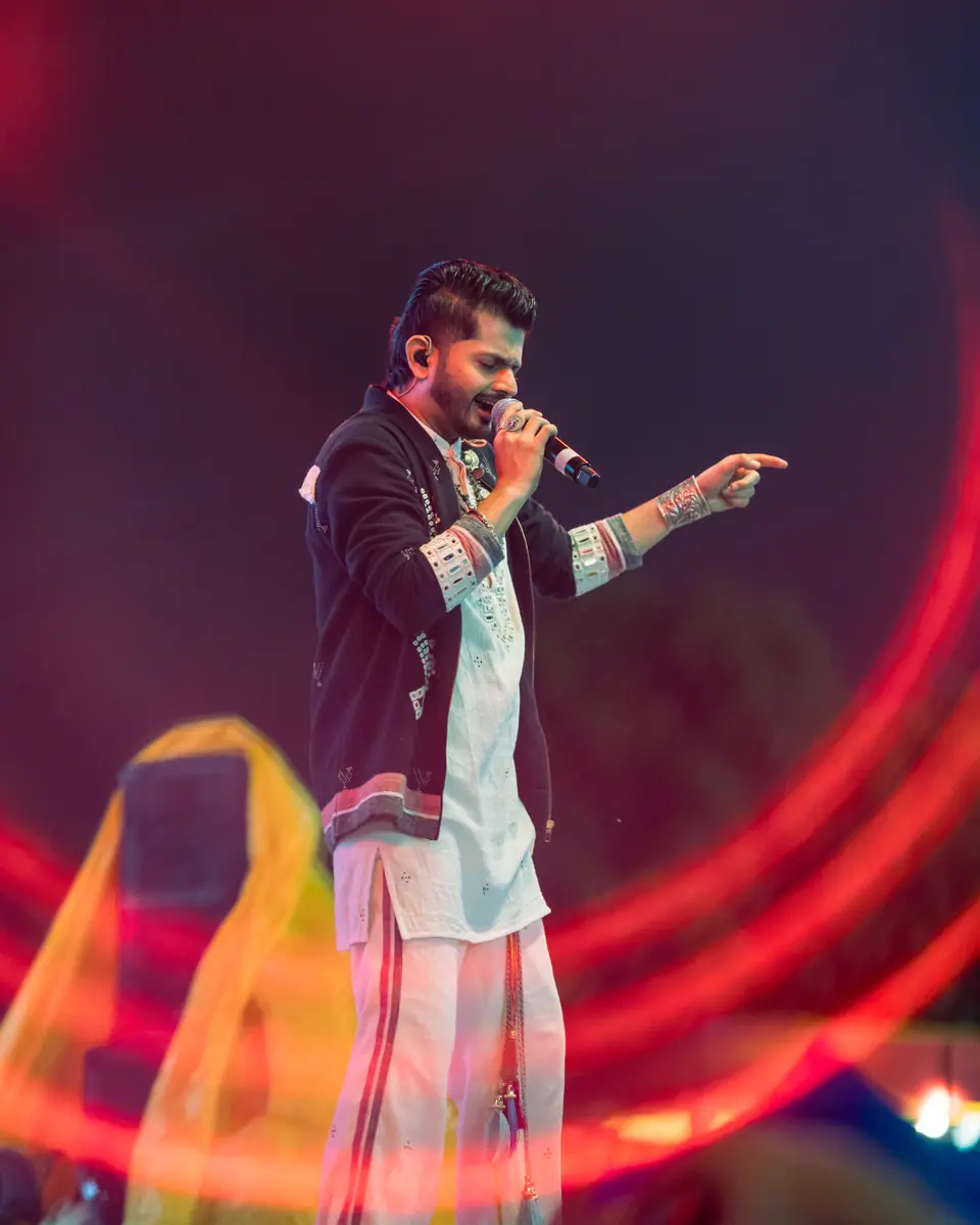 A singer in a black jacket over a white kurta, pointing at the crowd
